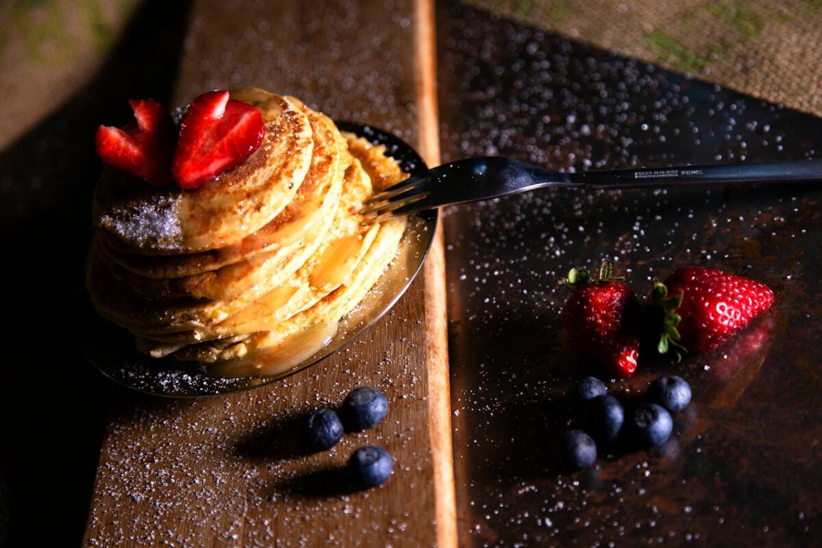 A stack of golden pancakes topped with strawberries, surrounded by fresh blueberries on a wooden board.