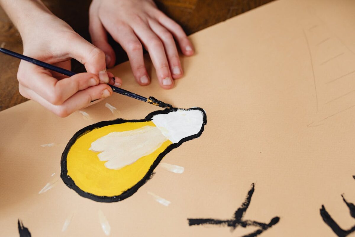A person paints a yellow lightbulb design on paper with a paintbrush, symbolizing creativity.